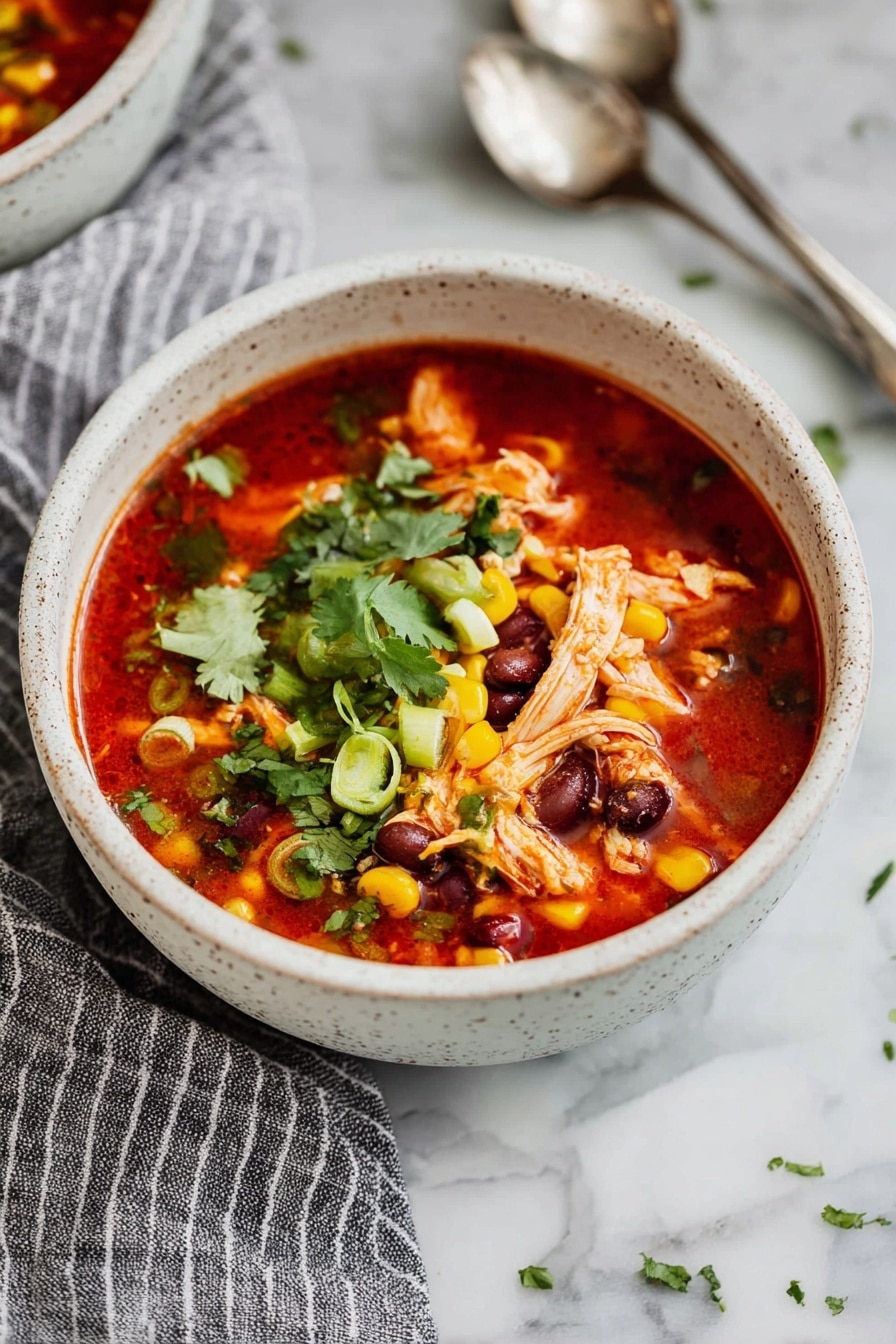 A white speckled bowl filled with a bright red soup, layered with shredded light brown chicken pieces, yellow corn kernels, and dark brown beans. The top layer is sprinkled with chopped green onions and fresh green cilantro leaves. The bowl sits on a white marbled surface with a grey and white striped cloth nearby and silver spoons to the side. photo taken with an iphone --ar 2:3 --v 7 - Instant Pot Southwest Chicken Soup, Southwest Chicken Soup recipe, quick chicken soup, easy Instant Pot recipes, hearty chicken soup