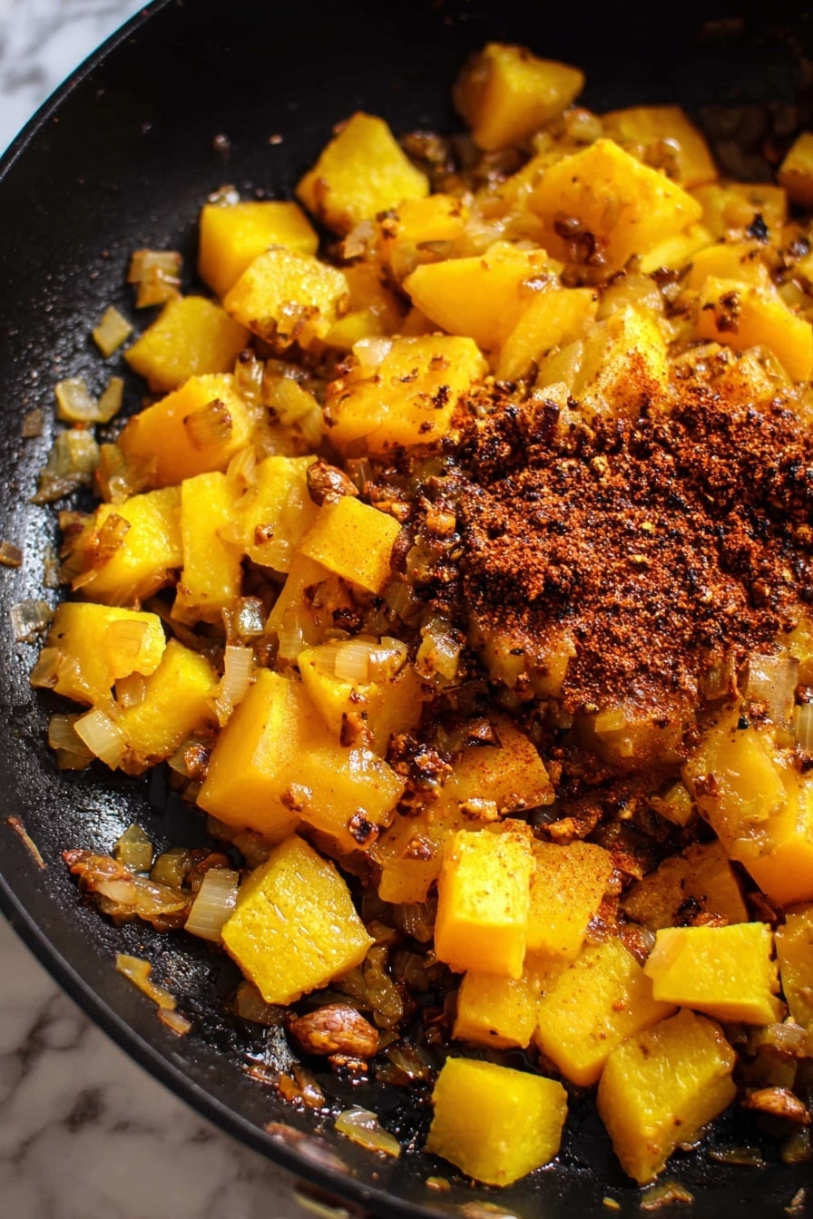 The image shows a black pan filled with cooked diced yellow squash and bits of cooked onion. There is a pile of reddish-brown spices and some minced garlic on top of the squash near the center. The squash pieces have a soft, slightly shiny texture with some browned edges, and the onions are translucent with some light browning. The black pan contrasts with the bright yellow and orange tones of the squash and the deep red of the spices. The background is a white marbled texture photo taken with an iphone --ar 2:3 --v 7 - Creamy Butternut Squash Butter Chicken, butternut squash chicken dinner, spicy coconut chicken, comfort food recipes, easy healthy chicken dinner