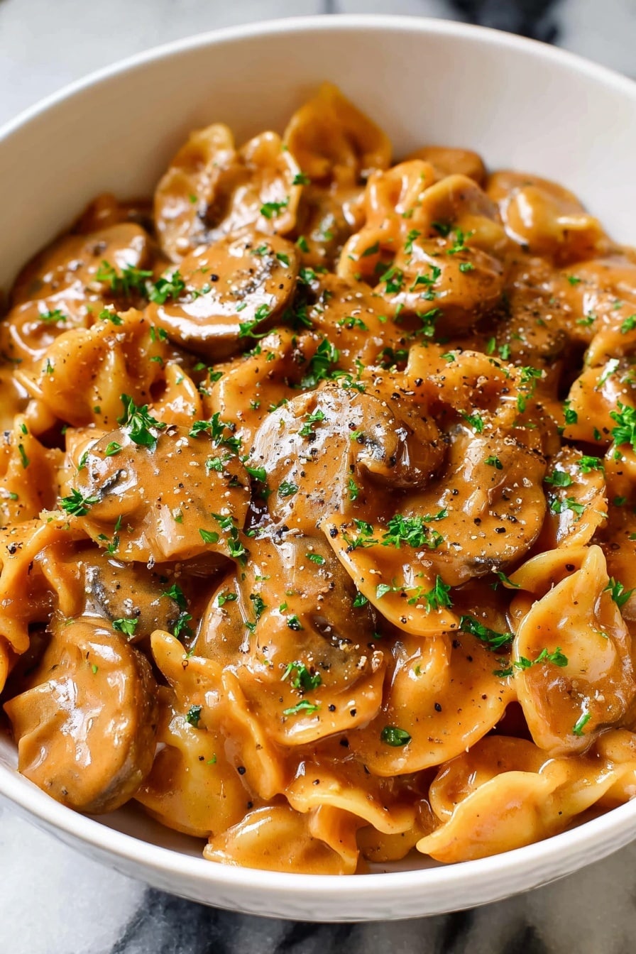A white bowl filled with creamy mushroom pasta, showing about two layers of plump, round mushroom slices and twisted pasta shapes fully coated in a rich, orange-brown sauce with a glossy texture. Small sprinkles of chopped green parsley and coarse black pepper are scattered evenly on top, giving a fresh contrast. The bowl sits on a white marbled surface. photo taken with an iphone --ar 2:3 --v 7 - French Onion Pasta with Caramelized Onions and Melted Gruyère, French onion pasta, caramelized onion pasta, cheesy pasta recipes, hearty pasta dishes
