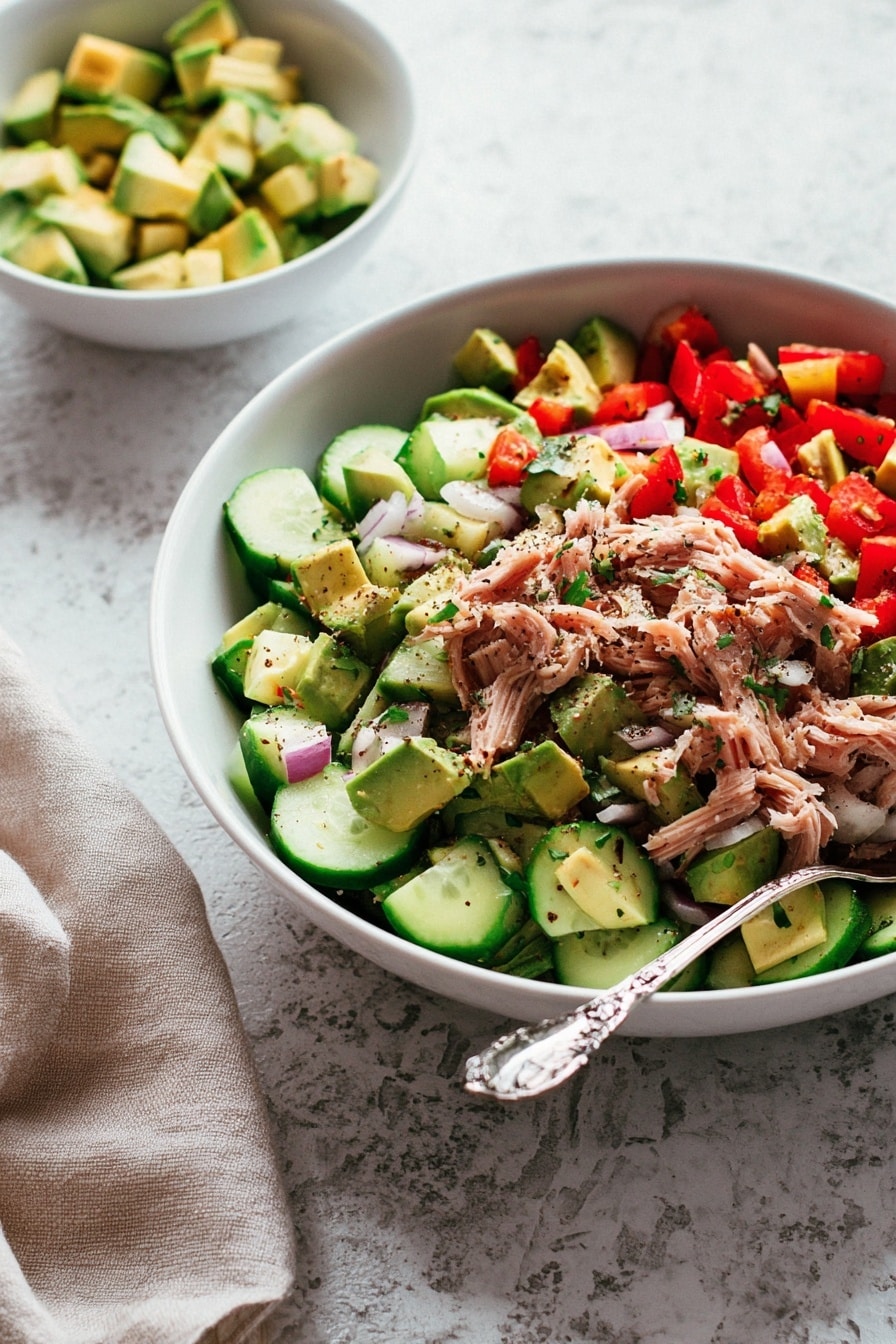 A white bowl filled with a colorful salad featuring three main layers: a base of bright green cucumber slices, a middle layer of light pink shredded meat, and a top layer of small diced red bell peppers, light purple onion pieces, and chunks of green avocado, all sprinkled lightly with black pepper and herbs. A detailed silver spoon rests inside the bowl on the right side. Behind the large bowl, there is a smaller white bowl with more diced avocado pieces. The dishes sit on a white marbled textured surface with a soft beige cloth napkin partially visible in the bottom left corner. The photo taken with an iphone --ar 2:3 --v 7 - Avocado Tuna Salad, healthy tuna salad, quick lunch recipes, easy seafood salad, fresh avocado salad