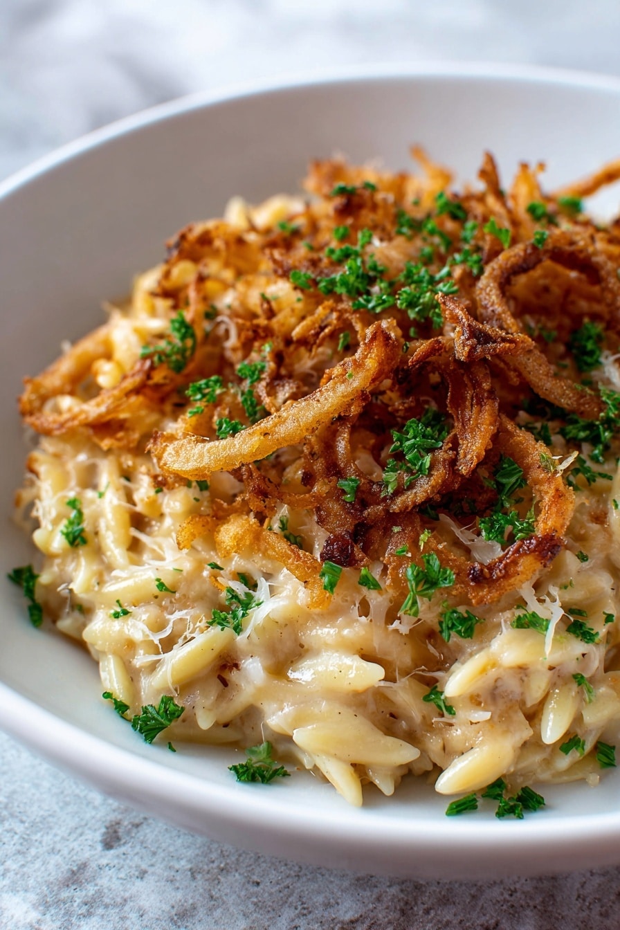 A close-up view of a pasta dish served in a white bowl. The bottom layer consists of small pasta pieces in a creamy, light beige sauce mixed with white shredded cheese. On top, there are golden brown crispy fried onion rings scattered evenly across the surface. Bright green chopped parsley is sprinkled over the entire dish for color contrast. The white bowl is placed on a white marbled textured surface. photo taken with an iphone --ar 2:3 --v 7 - French Onion Chicken Orzo Casserole, comforting chicken pasta bake, caramelized onion casserole, easy orzo casserole recipes, French onion-inspired dinner