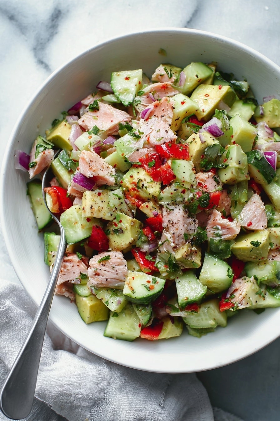 The image shows a white bowl filled with a colorful salad made of chopped layers. The base layer is light green cucumber pieces with dark green skin, mixed with cubed avocado of a soft green shade. Among these are scattered chunks of light pink cooked fish. Small pieces of red pepper and bits of light purple onion are spread throughout. The salad is lightly sprinkled with black pepper and fresh green herbs, giving a fresh look. The bowl is placed on a white marbled surface with part of a light gray cloth visible on the side. A metal spoon rests inside the bowl. Photo taken with an iphone --ar 2:3 --v 7 - Avocado Tuna Salad, healthy tuna salad, quick lunch recipes, easy seafood salad, fresh avocado salad