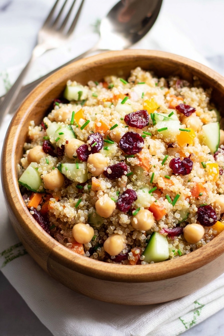 A wooden bowl filled with a colorful quinoa salad showing three main layers: the bottom layer is light beige quinoa grains, the middle layer has light tan chickpeas and small cubes of pale green cucumber and orange bell pepper, and the top layer is dotted with dark red dried cranberries and chopped bright green chives, all mixed together giving a textured and fresh look. The bowl is placed on a white marbled surface with a white cloth underneath and a silver fork and spoon to the side. Photo taken with an iphone --ar 2:3 --v 7 - Cranberry Apple Quinoa Salad, healthy Quinoa salad with cranberries and apples, easy nutritious Quinoa salad recipe, vibrant fruit and quinoa salad, quick quinoa side dish