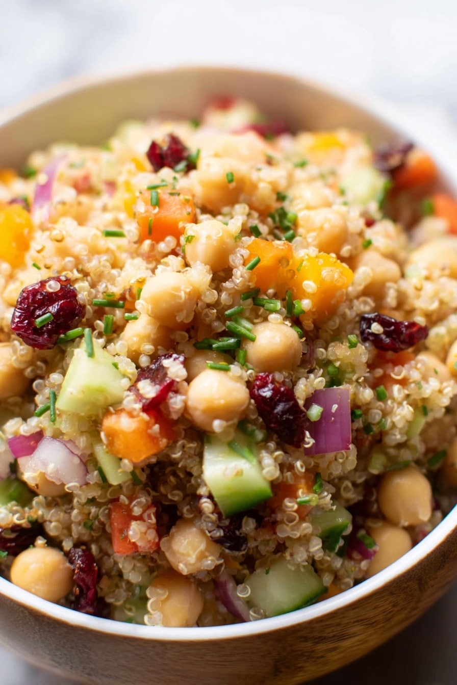 A close-up view of a bowl filled with a layered quinoa salad, showing tiny light tan quinoa grains as the base, mixed with layers of small chickpeas that are pale beige, bright orange bell pepper pieces, small cucumber chunks in light green, chopped red onions in light purple, dried dark red cranberries scattered evenly, and sprinkled green chopped chives on top. The bowl is white with a wooden exterior, sitting on a white marbled surface. The textures vary between soft quinoa, firm chickpeas, and crunchy vegetables, all mixed together in an inviting way. Photo taken with an iphone --ar 2:3 --v 7 - Cranberry Apple Quinoa Salad, healthy Quinoa salad with cranberries and apples, easy nutritious Quinoa salad recipe, vibrant fruit and quinoa salad, quick quinoa side dish