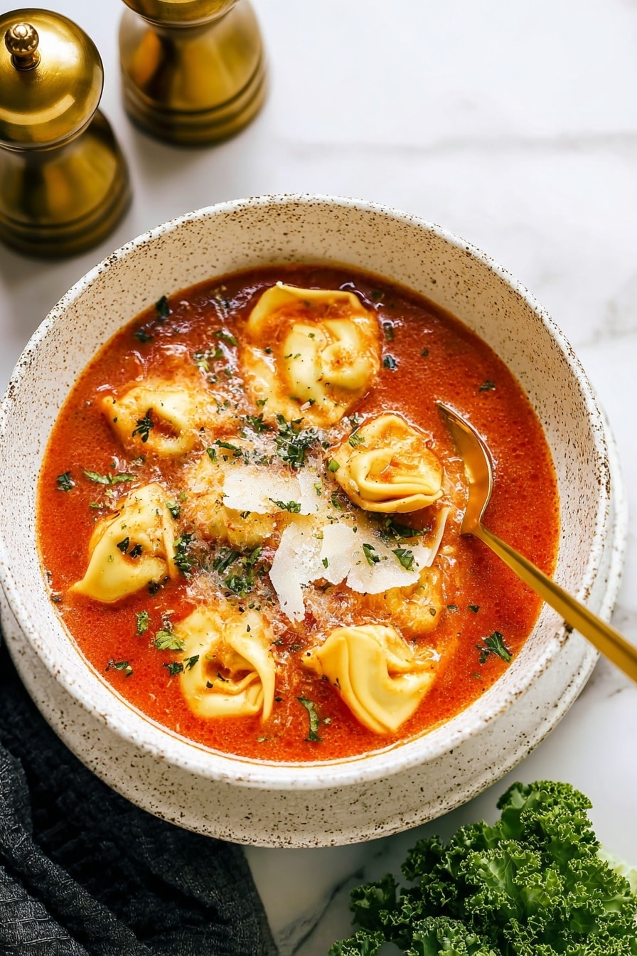 A white speckled bowl filled with bright red tomato soup, with about six plump yellow tortellini pasta floating on top. There are small green herbs sprinkled all over the soup and pasta. Thin white cheese shavings are scattered across the surface, melting slightly into the warm soup. A gold spoon rests in the bowl, partly submerged in the soup, on the right side. The bowl sits on a white marbled surface, with two brass salt and pepper mills blurred in the background on the upper left, and a piece of green kale partially visible on the lower right. The photo taken with an iphone --ar 2:3 --v 7 - Kale Tortellini Soup, kale tortellini soup recipe, hearty vegetable tortellini soup, quick kale tortellini dish, comforting pasta and greens soup