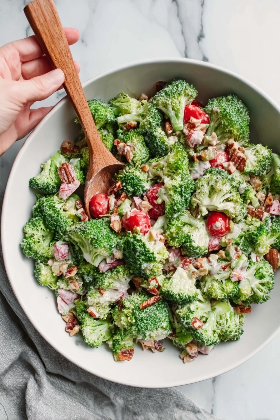 The image shows a white bowl filled with a fresh broccoli salad. The salad has bright green broccoli florets evenly spread throughout. Small red cherry tomatoes are mixed among the broccoli, adding pops of red color. There are also some light pink and white chunks, likely pieces of a creamy ingredient, scattered all over the bowl. Small bits of nuts or seeds add a crunchy texture and light brown color. A woman's hand is holding a wooden spoon mixing the salad. The bowl sits on a white marbled surface with a light gray cloth partially seen on the side. Photo taken with an iphone --ar 2:3 --v 7 - Broccoli Salad with Grapes and Bacon, crunchy broccoli salad, fruity bacon salad, homemade mayo dressing, healthy bacon salad