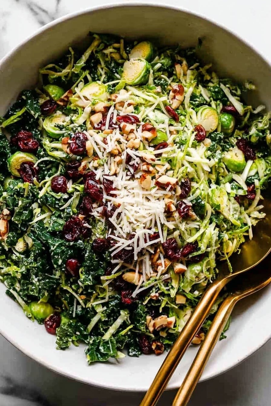 A large white bowl filled with a salad showing three main layers: the bottom layer is shredded dark green kale, the middle layer is thinly sliced bright green Brussels sprouts, and the top layer is sprinkled with small pieces of light brown chopped nuts, bright red dried cranberries, and thin white shredded cheese. A pair of dark golden spoons rests inside the bowl on the right side. The bowl is on a white marbled surface. Photo taken with an iphone --ar 2:3 --v 7 - Kale and Brussels Sprout Salad with Lemon Dijon Dressing and Toasted Nuts, healthy kale Brussels sprout salad, easy green salad recipe, citrus nut salad, nutritious vegetable salad