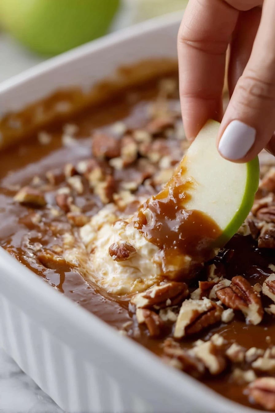 A close-up view of a white baking dish with a layered dessert. The bottom layer is light and creamy, topped with a smooth caramel layer that is dark brown and glossy. On top of the caramel, there are chopped pecans scattered around. A woman's hand is holding a green apple slice with a bite taken out of it, dipping into the creamy and caramel layers along with some pecans. The background surface is a white marbled texture. photo taken with an iphone --ar 2:3 --v 7 - Caramel Apple Cheesecake Dip, easy caramel apple dip, apple dessert dip, no-bake cheesecake dip, fall appetizer recipes