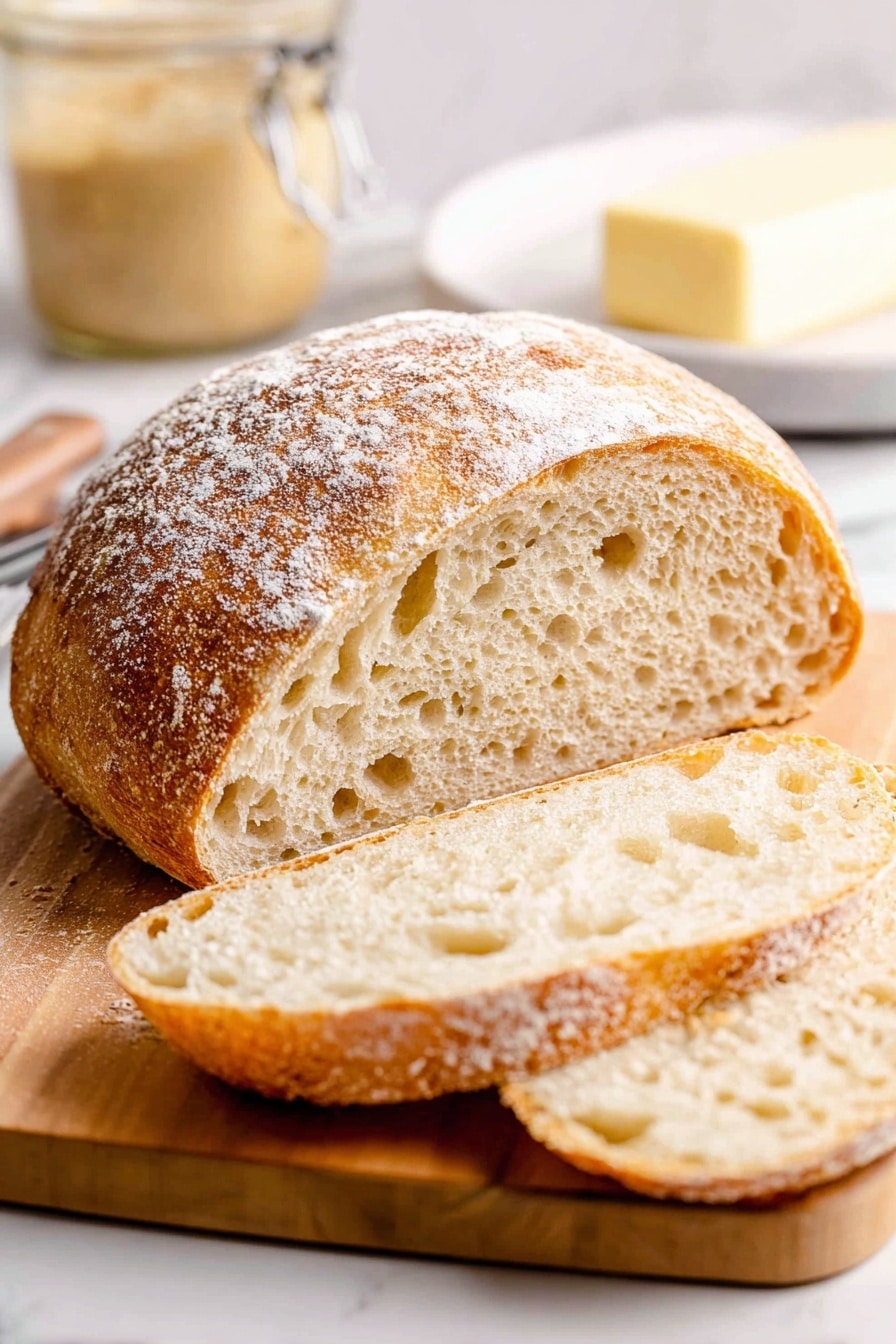 A round loaf of bread with a light dusting of flour on top sits on a wooden cutting board with a smooth light brown grain. In front of the loaf, there are two thick slices of bread showing a soft, airy inside with many small holes and a light golden crust. In the blurred background, there is a jar of creamy spread and a white dish with a stick of butter. The whole scene is set against a white marbled surface. photo taken with an iphone --ar 2:3 --v 7 - Homemade Sourdough Bread, sourdough bread recipe, how to make sourdough, artisanal bread, sourdough baking tips