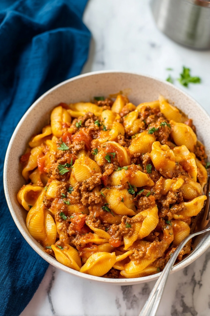 The image shows a white round bowl filled with three layers of food: the bottom layer is a small amount of sauce, the middle layer is cooked pasta shells in a bright orange color, and the top layer consists of crumbled brown meat mixed with bits of red tomatoes and green herbs scattered throughout. The bowl is placed on a white marbled surface with a dark blue cloth partially visible on the left side, and a blurry silver container is in the background to the right. The overall look is warm and hearty, with a close-up view highlighting the textures of pasta and meat. Photo taken with an iphone --ar 2:3 --v 7 - Instant Pot Taco Pasta, taco pasta recipe, easy instant pot dinners, quick pasta recipes, family-friendly taco pasta