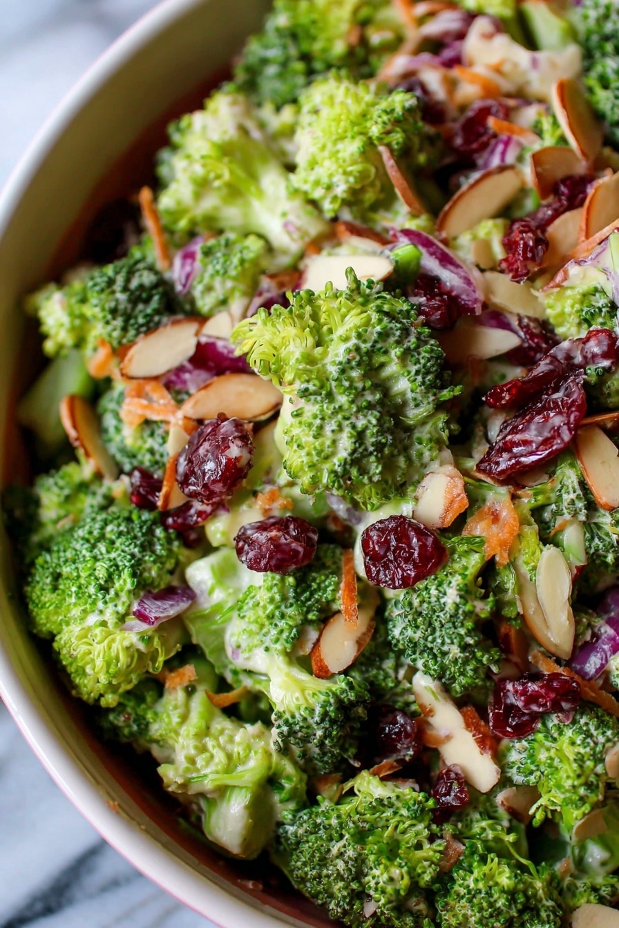 The image shows a close-up of a broccoli salad in a white bowl, placed on a white marbled surface. The salad has several layers with bright green broccoli florets, small pieces of red onion, deep red dried cranberries, thin almond slices, and a creamy dressing coating the ingredients. The broccoli pieces are the most visible and have a fresh, textured look. The red onion bits add hints of purple, and the dried cranberries bring pops of dark red color spread evenly. Thin beige almond slices are scattered throughout, adding contrast and texture. The creamy dressing covers some parts of the ingredients with a shiny, smooth surface. Photo taken with an iphone --ar 2:3 --v 7 - Broccoli Salad with Bacon, Cranberries, and Cheddar, broccoli salad with bacon, cranberry broccoli salad, cheesy broccoli salad, healthy broccoli salad, easy broccoli salad