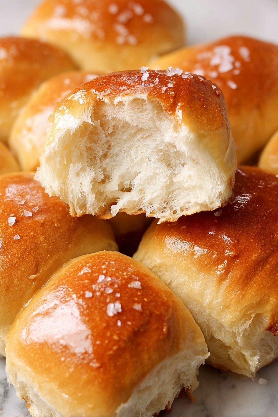 The image shows several golden-brown dinner rolls piled together inside a white bowl lined with white baking paper. Each roll has a smooth, shiny top with a slightly textured surface and is sprinkled with coarse salt crystals. The rolls have a soft and fluffy appearance with rounded edges, and their colors vary from light golden to deeper amber on the crust. The white marbled background is visible softly blurred behind the bowl, adding brightness to the scene. photo taken with an iphone --ar 2:3 --v 7 - Soft and Buttery Parker House Rolls, Parker House Rolls, How to Make Parker House Rolls, Easy Parker House Rolls, Homemade Parker House Rolls
