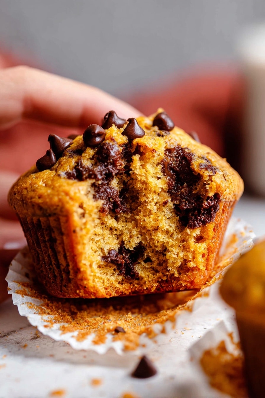 A close-up picture shows a chocolate chip muffin torn in half, held by a woman's hand on the left. The muffin has a warm golden-brown color with soft, moist texture inside. Big dark brown melted chocolate chips are spread throughout the muffin, some sitting on the top, some inside. The muffin liner is white and sits on a white marbled surface with crumbs scattered around. The background is blurry with a mix of soft gray and reddish hues. photo taken with an iphone --ar 2:3 --v 7 - Pumpkin Chocolate Chip Muffins, pumpkin muffin recipes, fall dessert ideas, easy pumpkin muffins, chocolate chip muffin recipe