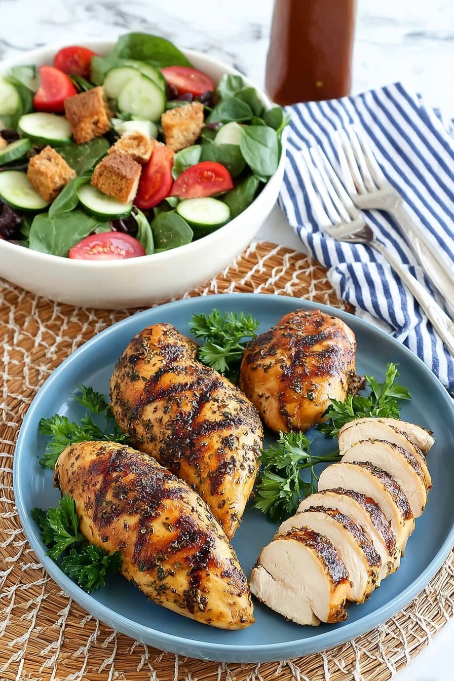 The image shows a dish with four grilled chicken pieces on a blue plate placed on a woven mat with a white marbled surface underneath. Three of the chicken pieces are whole, golden brown with grill marks and dark herbs visible on the skin, each resting on fresh green parsley leaves scattered on the plate. The fourth chicken piece is sliced into about nine pieces, showing tender white inside with similar grill marks on the outside. Behind the plate, there is a white bowl filled with a fresh salad containing dark green spinach and lettuce leaves, bright red tomato wedges, round cucumber slices, and brown croutons. A striped blue and white napkin with two silver forks lies next to the plate, and a brown sauce bottle is slightly visible in the background. Photo taken with an iphone --ar 2:3 --v 7 - Air Fryer Lemon Herb Chicken Breasts, lemon herb chicken, healthy chicken recipes, quick chicken dinner, easy air fryer chicken