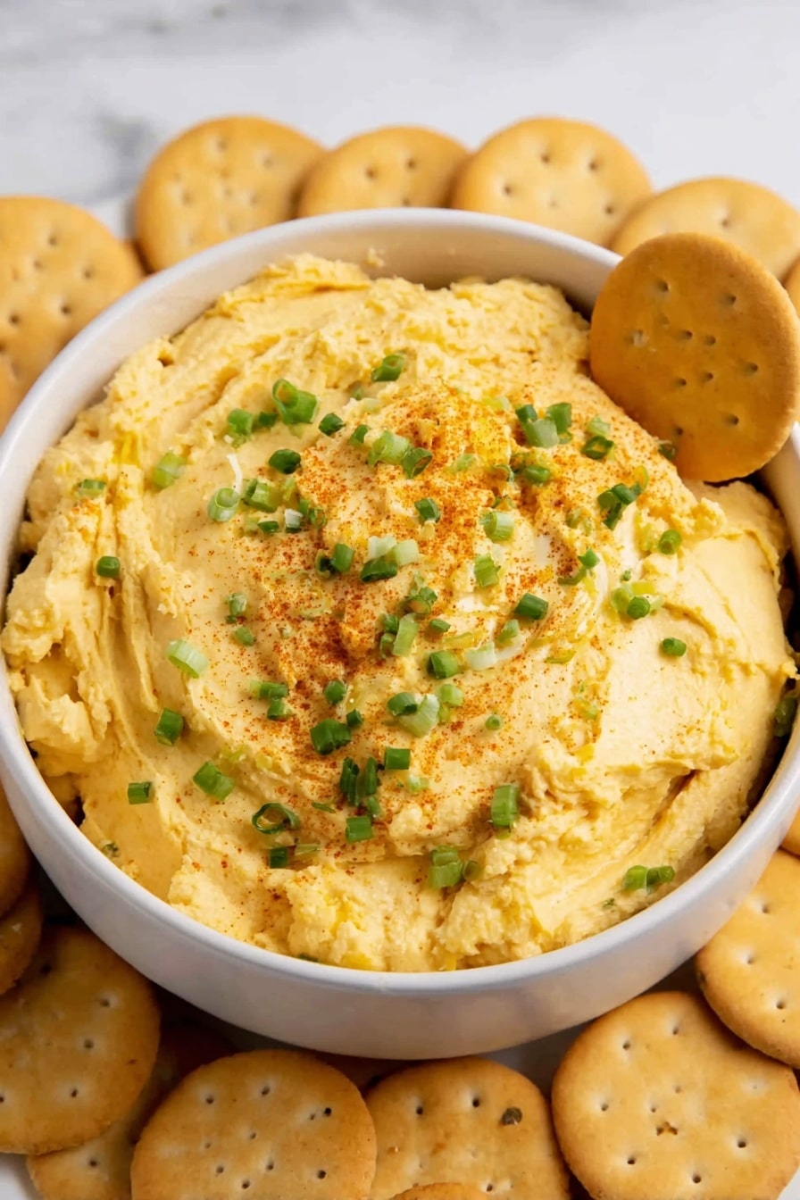 A white bowl is filled with a thick, yellowish spread that looks creamy and soft, with rough swirls and small bits of green herbs mixed inside. On top of the spread, there are light green chopped pieces and reddish brown powder sprinkled lightly. Several round, pale brown crackers with little holes are partially stuck into the spread and scattered around the bowl on a white marbled surface. The contrast between the yellow spread, the green herbs, and the brown crackers creates a warm, inviting look. photo taken with an iphone --ar 2:3 --v 7 - Deviled Eggs Dip, easy deviled eggs dip, creamy egg dip, party appetizer dip, quick snack recipe
