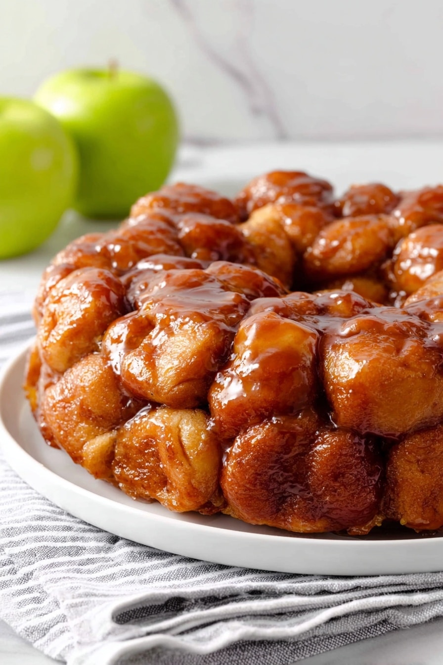 A round monkey bread made of many small, soft dough pieces coated in shiny caramel glaze, creating a glossy, sticky texture that covers the entire top and sides. The dough pieces are golden brown with a visible cinnamon-spiced sugar coating. The bread is placed on a white plate with a white marbled surface beneath it, and a green apple is blurred in the background along with stacked white plates on the right. photo taken with an iphone --ar 2:3 --v 7 - Caramel Apple Monkey Bread, apple monkey bread, caramel apple dessert, easy fall dessert, printable monkey bread recipe