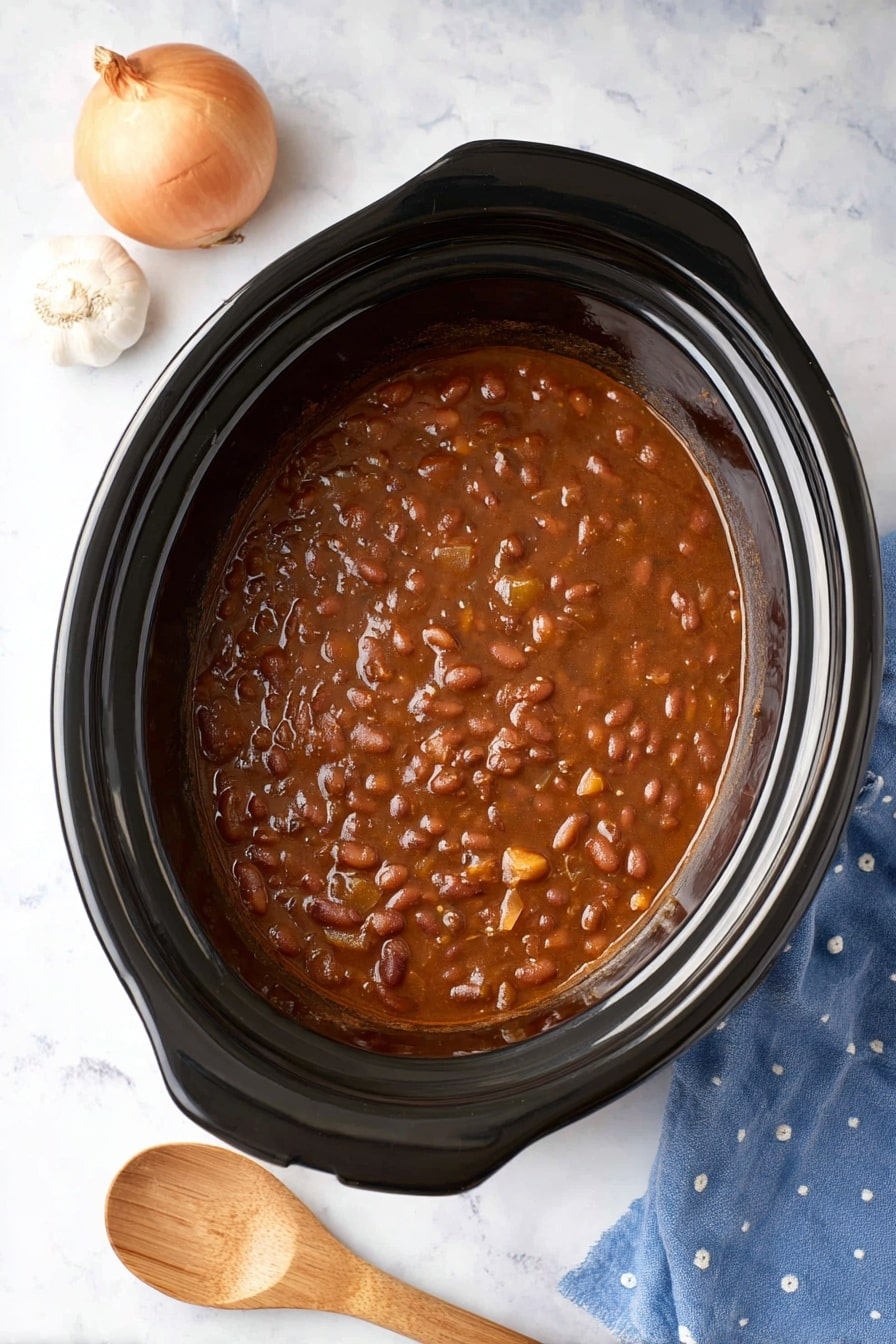A top-down view of a black slow cooker filled with thick brown bean stew with visible beans and small bits of vegetables mixed evenly throughout. The stew fills most of the cooker, showing a glossy texture on top. To the top left of the cooker, a whole onion with skin and a small bulb of garlic rest on a white marbled surface. At the bottom right corner, part of a blue cloth with white dots is visible next to a wooden spoon with a smooth light wood grain pattern. photo taken with an iphone --ar 2:3 --v 7 - Classic BBQ Slow Cooker Baked Beans, BBQ baked beans, slow cooker baked beans, smoky BBQ side dish, easy baked beans recipe