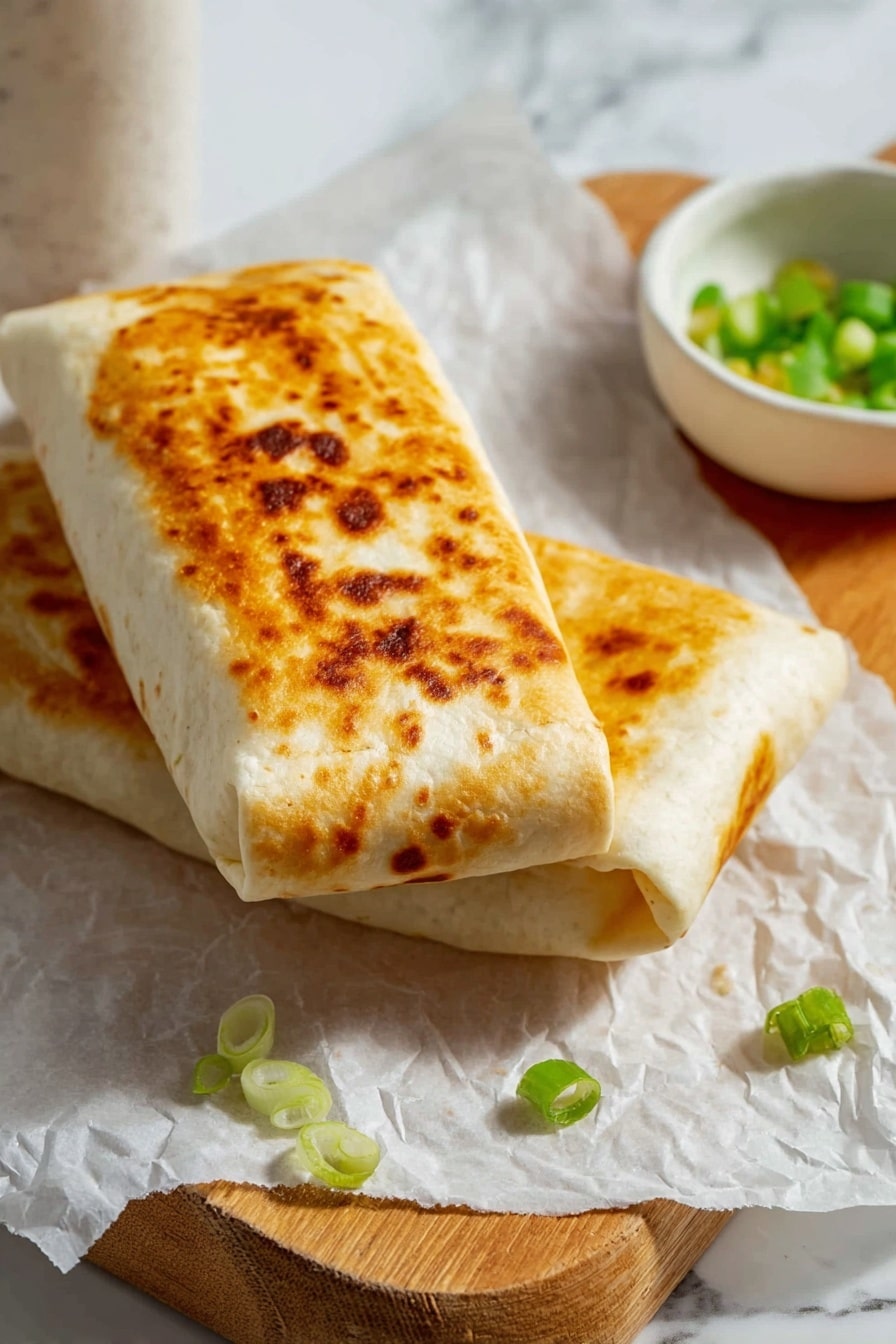Two rolled burritos lie on white parchment paper on a wooden board. The top burrito has a golden brown crisp texture on the outside with some darker spots, showing it has been toasted. The second burrito is tucked slightly underneath, showing the smooth, pale tortilla wrap. Small pieces of green onion are scattered around, and a small white ceramic bowl holding chopped green onions is nearby. The background is a white marbled texture. Photo taken with an iphone --ar 2:3 --v 7 - Cheesy Garlic Chicken Wraps, easy chicken wraps, quick dinner ideas, garlic chicken wraps, cheesy lunch recipes