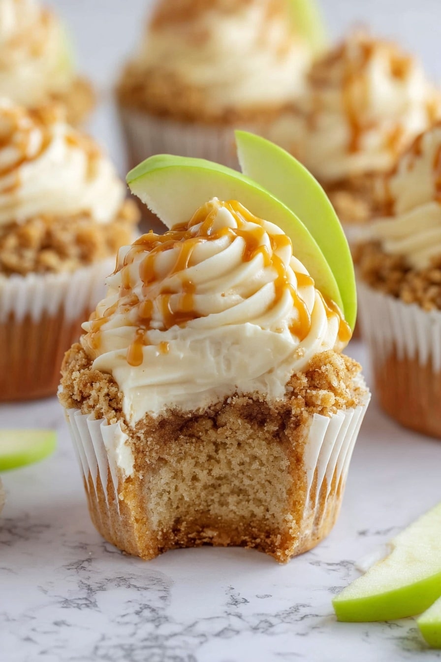 The image shows a close-up of a cupcake with its white wrapper peeled down around the base, revealing a light brown crumb with a soft texture. The cupcake is topped with a crumbly oat mixture layer that looks golden and slightly crunchy. Above this is a thick swirl of creamy off-white frosting with smooth peaks, drizzled with a thin orange sauce. A fresh green apple slice is placed leaning against the frosting on top. In the background, several similar cupcakes are softly focused, set on a white marbled surface, with a few green apple slices scattered nearby. photo taken with an iphone --ar 2:3 --v 7 - Apple Crumble Cupcakes, fall apple cupcakes, easy apple dessert, cinnamon apple cupcakes, delicious apple crumble treats