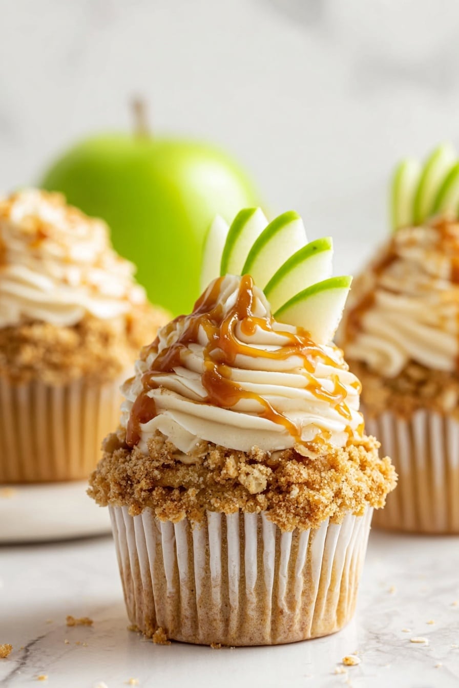 The image shows a close-up of a cupcake with three main layers. The bottom layer is a golden-brown cupcake wrapped in a white paper liner. On top of the cupcake is a crumbly, crunchy oat topping with light brown color and some oat flakes visible. The next layer is a swirl of light beige frosting with a smooth, creamy texture. This frosting is decorated with a thin drizzle of caramel sauce, adding glossy orange-brown streaks. A fresh green apple slice is placed upright at the back of the frosting swirl, adding a fresh look with its bright green color. The cupcake is placed on a smooth white marble surface, with two more similar cupcakes blurred in the background and a bright green apple out of focus. Photo taken with an iphone --ar 2:3 --v 7 - Apple Crumble Cupcakes, fall apple cupcakes, easy apple dessert, cinnamon apple cupcakes, delicious apple crumble treats