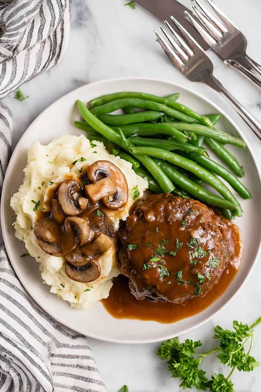 A white plate holds three main parts: on the left, a creamy heap of mashed potatoes topped with brown mushroom gravy and whole mushroom slices; on the top, a neat bunch of bright green steamed green beans; on the right, a large round meat patty covered thickly with shiny brown mushroom gravy and sprinkled with small green herbs. The plate is set on a white marbled surface with a striped cloth and silver fork and knife in the blurry background. A few fresh green parsley leaves are on the surface near the plate. Photo taken with an iphone --ar 2:3 --v 7 - Homestyle Salisbury Steaks in Rich Mushroom Gravy, Salisbury Steaks recipe, mushroom gravy recipe, comforting beef dinner, hearty homemade Salisbury Steaks
