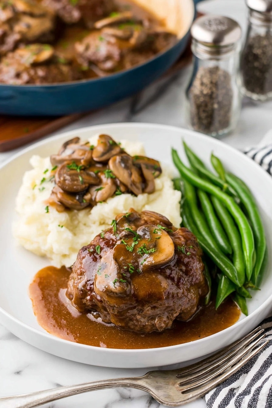 A white plate is filled with three main parts: a large brown meat patty covered with a glossy brown mushroom gravy sauce sprinkled with small green herbs on top, a mound of creamy white mashed potatoes topped with the same mushroom gravy and visible sliced brown mushrooms, and a neat row of bright green cooked green beans placed on the side. The plate rests on a white marbled surface, with a blue pan filled with more brown meat patties and mushroom sauce blurred in the background, along with a salt and pepper shaker. A black and white striped cloth with a silver fork is placed on the bottom right corner. photo taken with an iphone --ar 2:3 --v 7 - Homestyle Salisbury Steaks in Rich Mushroom Gravy, Salisbury Steaks recipe, mushroom gravy recipe, comforting beef dinner, hearty homemade Salisbury Steaks