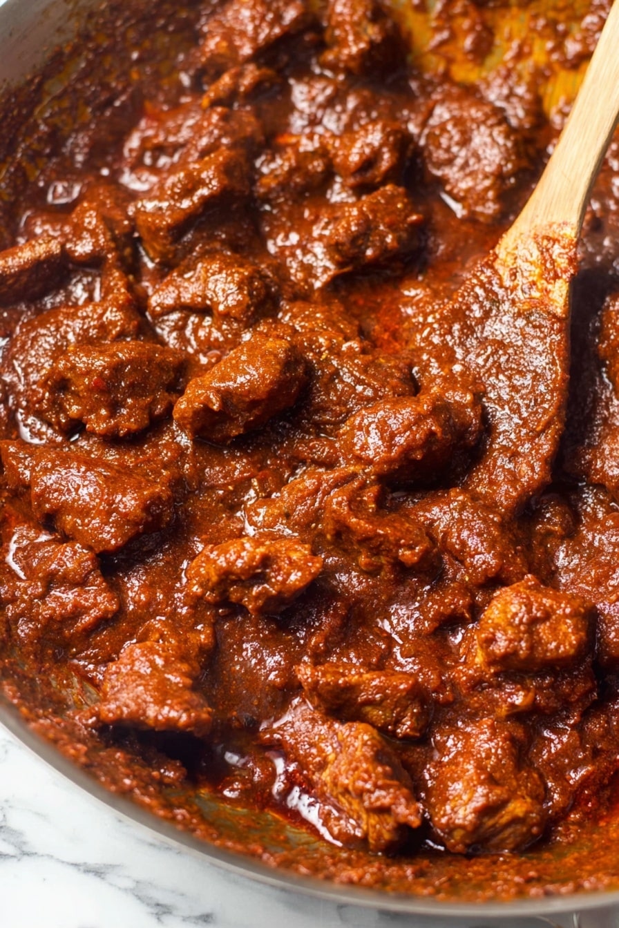 The image shows a close-up of many small, soft pieces of meat covered in a thick, rich reddish-brown sauce in a metal pan. The sauce has a slightly chunky and glossy texture, sticking well to the meat pieces. A wooden spoon is partly visible in the upper right corner, mixing the meat and sauce. The pan’s edges have traces of the sauce, giving a rough contrast to the main dish. The background is a white marbled texture. photo taken with an iphone --ar 2:3 --v 7 - Hearty Carne Guisada, Carne Guisada with Tender Beef, Spiced Tomato Gravy Recipe, Slow Cooker Carne Guisada, Comforting Beef Stew Recipe