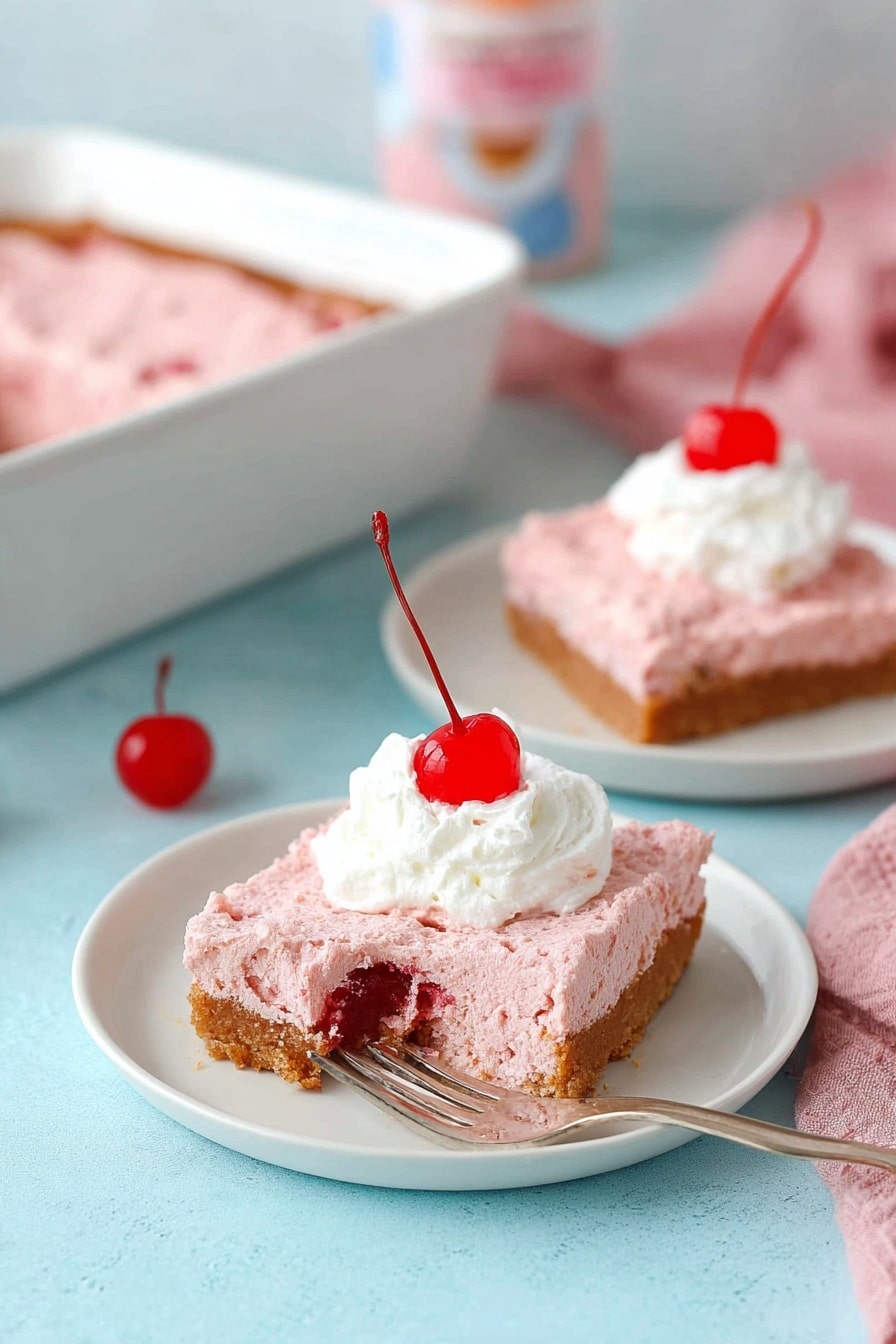 The image shows a slice of pink cake with visible red fruit pieces inside, placed on a small white plate. The cake has two main layers with a light, soft texture and a slightly browned edge on top. On top of the cake slice, there is a swirl of white whipped cream with a bright red cherry placed in the center. A silver fork rests on the right side of the plate. In the background, there is another slice of the same cake on a white plate, slightly blurred, and a few containers out of focus. The setting has a clean, bright look with a white marbled surface. Photo taken with an iphone --ar 2:3 --v 7 - 2-Ingredient Cherry Angel Food Cake, cherry angel food cake, easy cherry dessert, 2-ingredient cake recipe, no-bake cherry cake