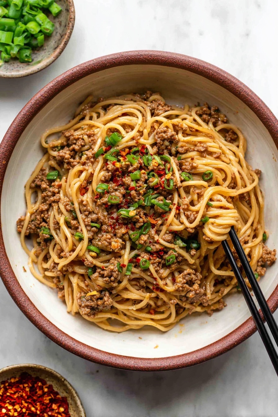 Mongolian Ground Beef and Udon Noodles Recipe 5 The dish is a bowl of noodles mixed with ground meat and small bits of green onions scattered on top. The noodles are light brown and thin, with the meat appearing darker brown and crumbly, evenly spread throughout. Small red chili flakes are sprinkled over the dish, adding some color contrast. Black chopsticks rest inside the bowl, picking up some noodles from the right side. The bowl is white with a rustic brown inside and sits on a white marbled surface. Two small bowls are partially visible in the background, holding chopped green onions and red chili flakes. Photo taken with an iphone --ar 2:3 --v 7 - Mongolian Ground Beef and Udon Noodles, Mongolian Beef Udon Stir-fry, Quick Asian Ground Beef Noodle Bowl, Savory Udon Noodles with Ground Beef, Easy Mongolian Beef and Udon Recipe