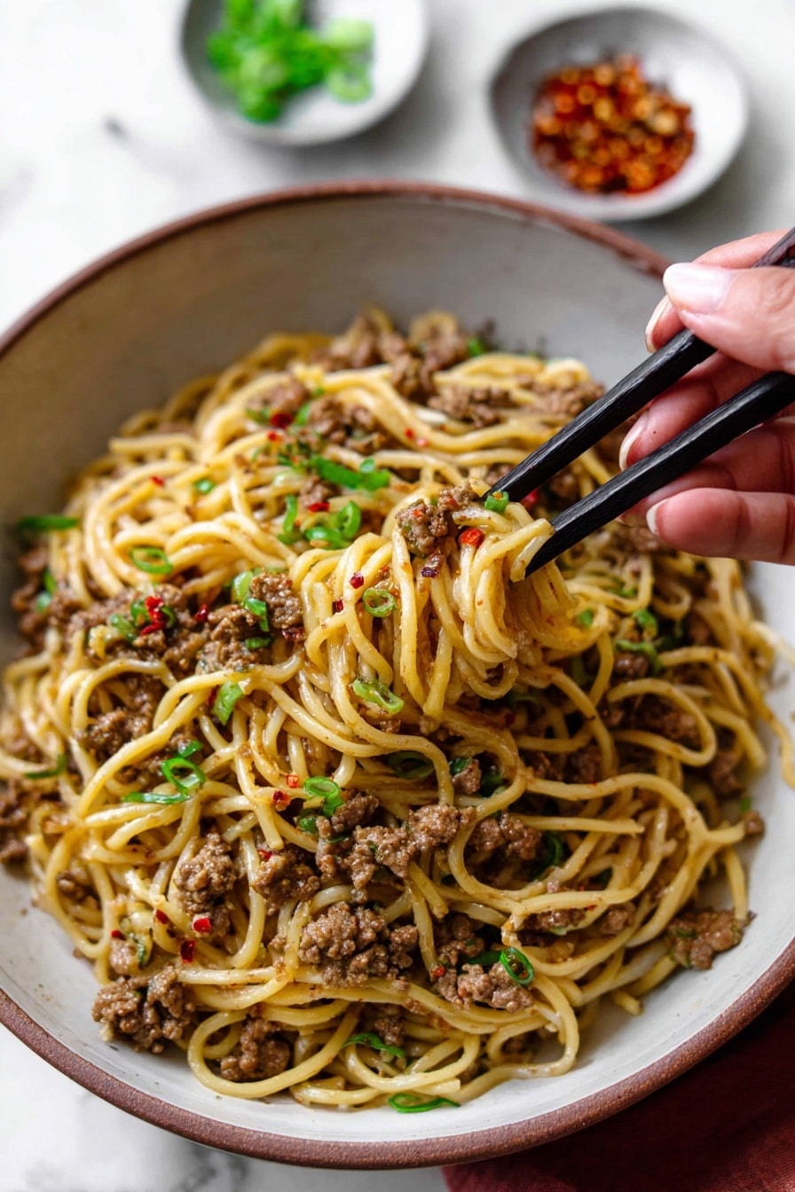 A close-up view of a large white bowl filled with one layer of cooked yellowish noodles mixed with small pieces of brown cooked ground meat, scattered with small bits of red chili flakes and green sliced scallions on top. A woman's hand holding black chopsticks is lifting a small bundle of noodles and meat from the right side of the bowl. In the blurred background, there are two small white bowls, one with green sliced scallions and the other with crushed red pepper. The bowl is placed on a white marbled surface. Photo taken with an iphone --ar 2:3 --v 7 - Mongolian Ground Beef and Noodles, Mongolian beef and noodles, easy Mongolian beef recipe, quick beef and noodle stir-fry, savory ground beef noodles
