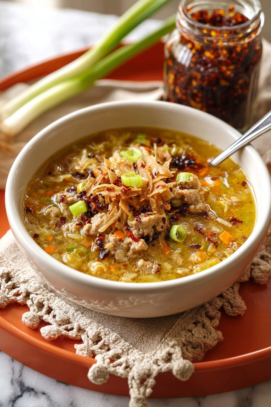 A white bowl filled with a thick soup sits on a beige cloth with crocheted edges on an orange tray. The soup has several layers: the bottom layer is a golden broth mixed with small chunks of light brown cooked meat and bits of soft, light yellow and orange vegetables. On top, there are scattered pieces of thin, crispy, light brown strips. The soup is garnished with small bright green sliced scallions and a sprinkle of red chili flakes, adding a pop of color. In the background, there is a glass jar with chili paste and some green onion stalks resting on the white marbled surface. Photo taken with an iphone --ar 2:3 --v 7 - Egg Roll Soup, Egg Roll Soup recipe, Asian-inspired soup, quick dinner ideas, easy egg roll soup