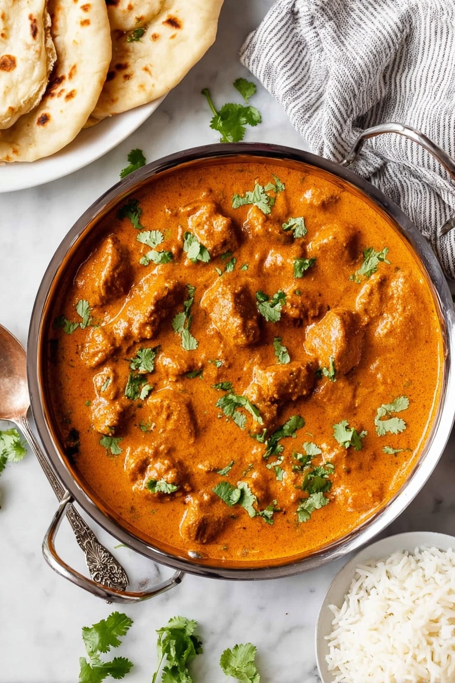 A shallow stainless steel pan filled with a thick, creamy orange curry sauce covering many chunks of tender meat, topped with scattered fresh green cilantro leaves. The pan rests on a white marbled surface with some cilantro leaves around it. To the top left, a white plate holds several pieces of lightly browned flatbread over a gray and white striped cloth. To the bottom center, a white bowl contains fluffy white rice. A silver spoon with an ornate handle lies on the left side near the pan. The bright colors and textures show a rich and hearty meal. photo taken with an iphone --ar 2:3 --v 7 - Creamy Chicken Tikka Masala, Chicken Tikka Masala recipe, Indian chicken curry, flavorful chicken curry, easy tikka masala recipe