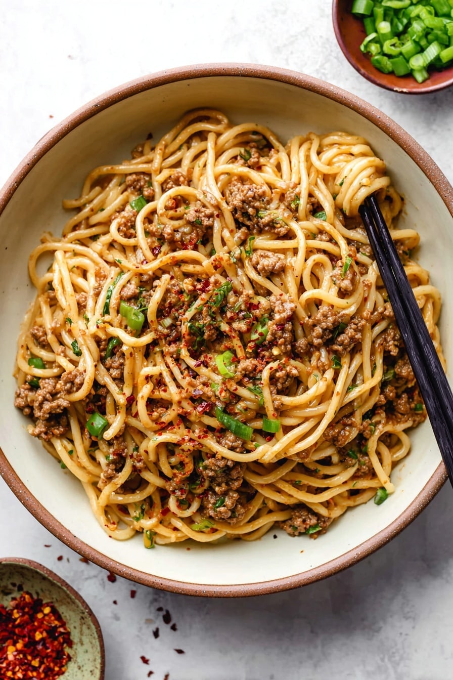 A round white bowl filled with a single layer of cooked noodles mixed with browned minced meat, scattered green onion pieces, and red pepper flakes on top. The noodles are light brown, coated with sauce, and twirled around a pair of black chopsticks placed on the right side inside the bowl. The background is a white marbled texture with small bowls of green onions and red pepper flakes partially visible near the top right corner. Photo taken with an iphone --ar 2:3 --v 7 - Mongolian Ground Beef and Noodles, Mongolian beef and noodles, easy Mongolian beef recipe, quick beef and noodle stir-fry, savory ground beef noodles