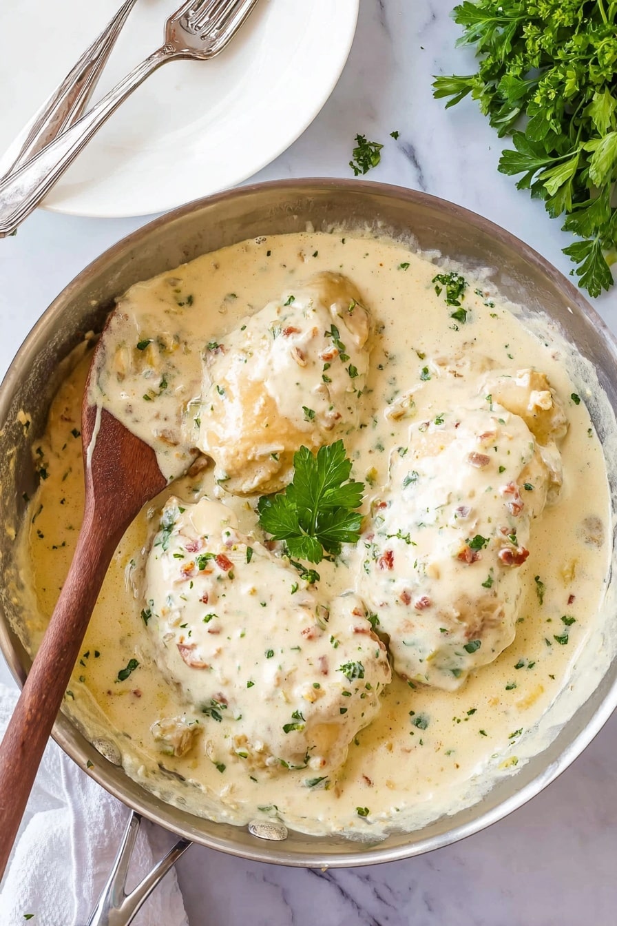 The image shows a metal pan filled with three large chicken pieces covered in a thick, creamy white sauce with small green herb bits and tiny reddish pieces scattered throughout. The sauce has a smooth, rich texture that coats the chicken evenly. A wooden spoon rests on the left side of the pan, partially covered in sauce. In the center of the dish, a sprig of fresh green parsley adds color contrast. The pan sits on a white marbled surface with a white plate holding two forks in the upper left corner and some green parsley leaves visible on the top right side of the frame. Photo taken with an iphone --ar 2:3 --v 7 - Creamy Smothered Chicken, creamy chicken dinner, chicken with mushroom sauce, easy comfort food recipes, crispy bacon chicken