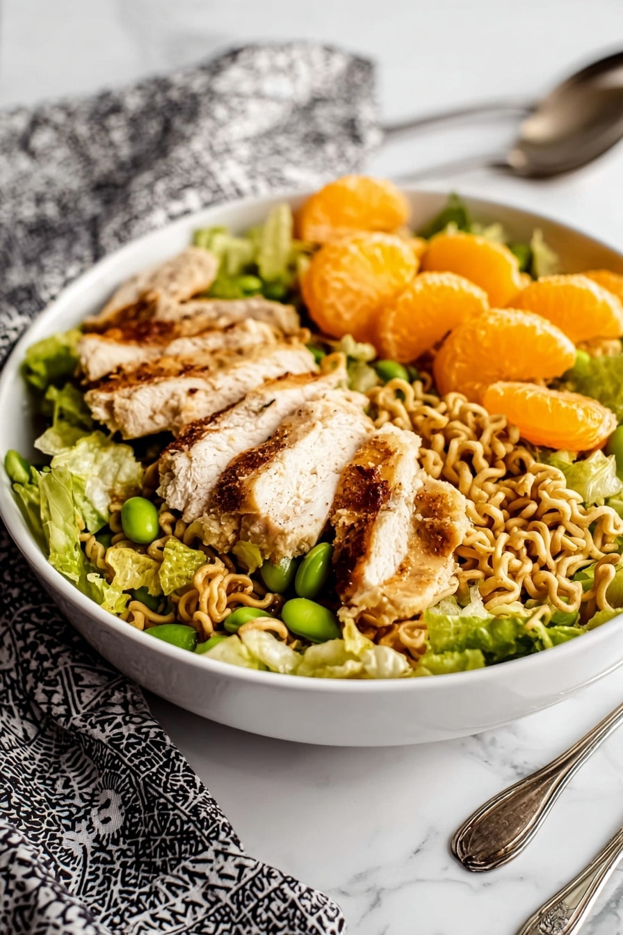 A white bowl filled with a layered salad starting with a base of green leafy lettuce, topped with light brown cooked noodles that appear wavy and curly. On top of the noodles are several slices of grilled chicken with a golden brown outer layer and white inside, scattered evenly. Bright orange mandarin slices are placed above the chicken and noodles, along with small green edamame beans dispersed throughout the salad. The bowl sits on a white marbled surface with a folded black and white patterned cloth in the background, and vintage silver fork and spoon resting near the bowl. Photo taken with an iphone --ar 2:3 --v 7 - Chinese Chicken Salad, Chinese Chicken Salad recipe, healthy Chinese chicken salad, easy Chinese salad, crunchy chicken salad