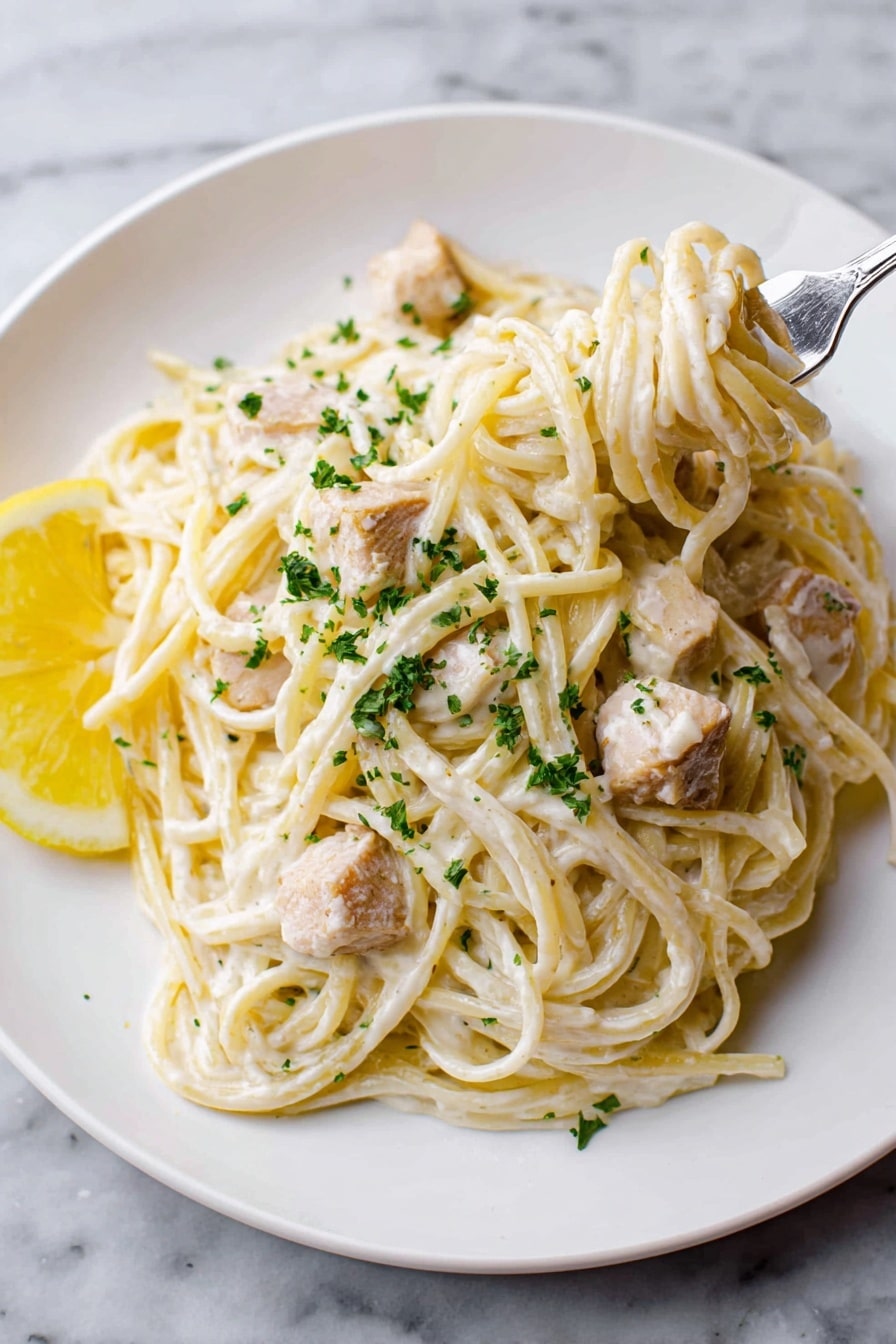 Creamy Lemon Chicken Pasta Recipe 7 A white plate holds a creamy pasta dish with about three visible layers of spaghetti coated in a thick white sauce. Scattered across and mixed in the pasta are small light brown cubes of chicken. There is a lemon slice placed on the left side of the plate, and green parsley flakes sprinkled on top to add color. A silver fork lifts a small twirl of pasta from the right side of the plate. The background is a white marbled surface. Photo taken with an iphone --ar 2:3 --v 7 - Creamy Lemon Chicken Pasta, Lemon Chicken Pasta, Easy Lemon Chicken Dinner, Bright Creamy Pasta, Quick Chicken Pasta