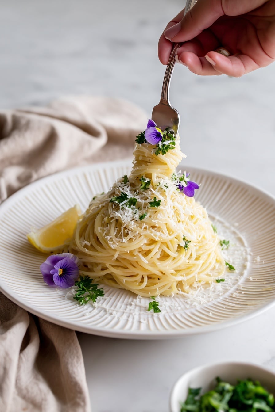 A white plate with thin ridges around the edge holds a mound of cooked spaghetti, pale yellow in color and loosely twisted. On top, finely grated cheese is sprinkled lightly, with fresh green parsley leaves scattered over, adding a bright contrast. Two small purple flowers and a thin lemon slice rest on one side of the pasta mound, giving a fresh and delicate touch. In the background, a beige cloth napkin is draped beside the plate on a white marbled surface. A small white bowl with chopped greens and lemon wedges is partly visible at the lower right corner. Photo taken with an iphone --ar 2:3 --v 7 - One Pot Lemon Pasta, Lemon Pasta Recipe, Easy Lemon Pasta, Quick Pasta Dinner, Fresh Lemon Pasta