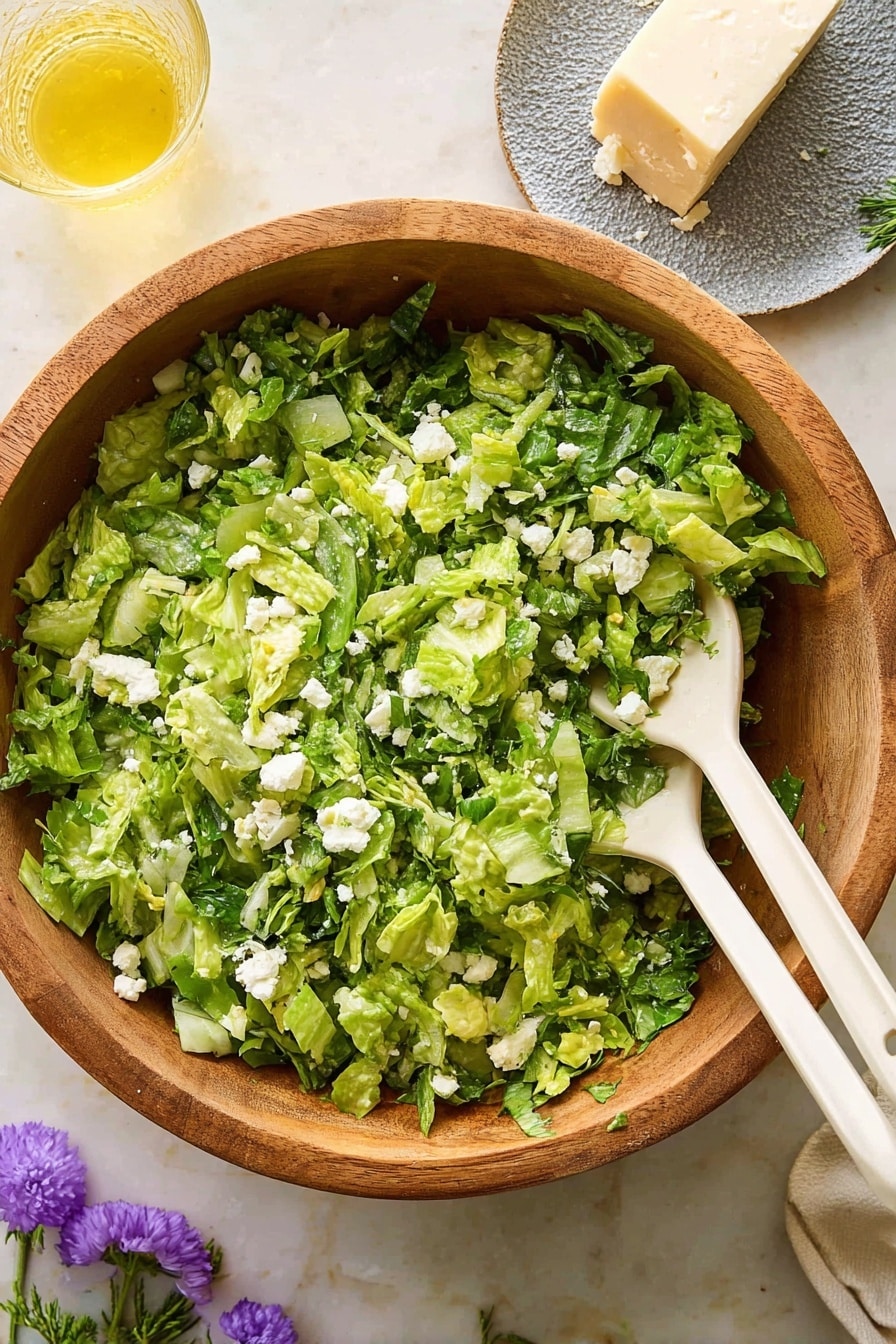 Greek Lettuce Salad with Feta, Dill, and Lemon Recipe 5 A wooden bowl holds a fresh green salad made of torn lettuce and small pieces of white cheese, mixed with finely chopped herbs like dill. Two white plastic salad spoons rest inside the bowl, partly covered by the leafy greens. To the right, there is a white plate with a heart-shaped piece of white cheese. The setting is on a white marbled textured surface, with a glass of light yellow liquid visible in the top left corner, and small purple and white flowers decorating the background. Photo taken with an iphone --ar 2:3 --v 7 - Greek Lettuce Salad with Feta, Dill, and Lemon, Mediterranean Salad, Easy Greek Salad Recipe, Fresh Lettuce Salad, Summer Salad Ideas